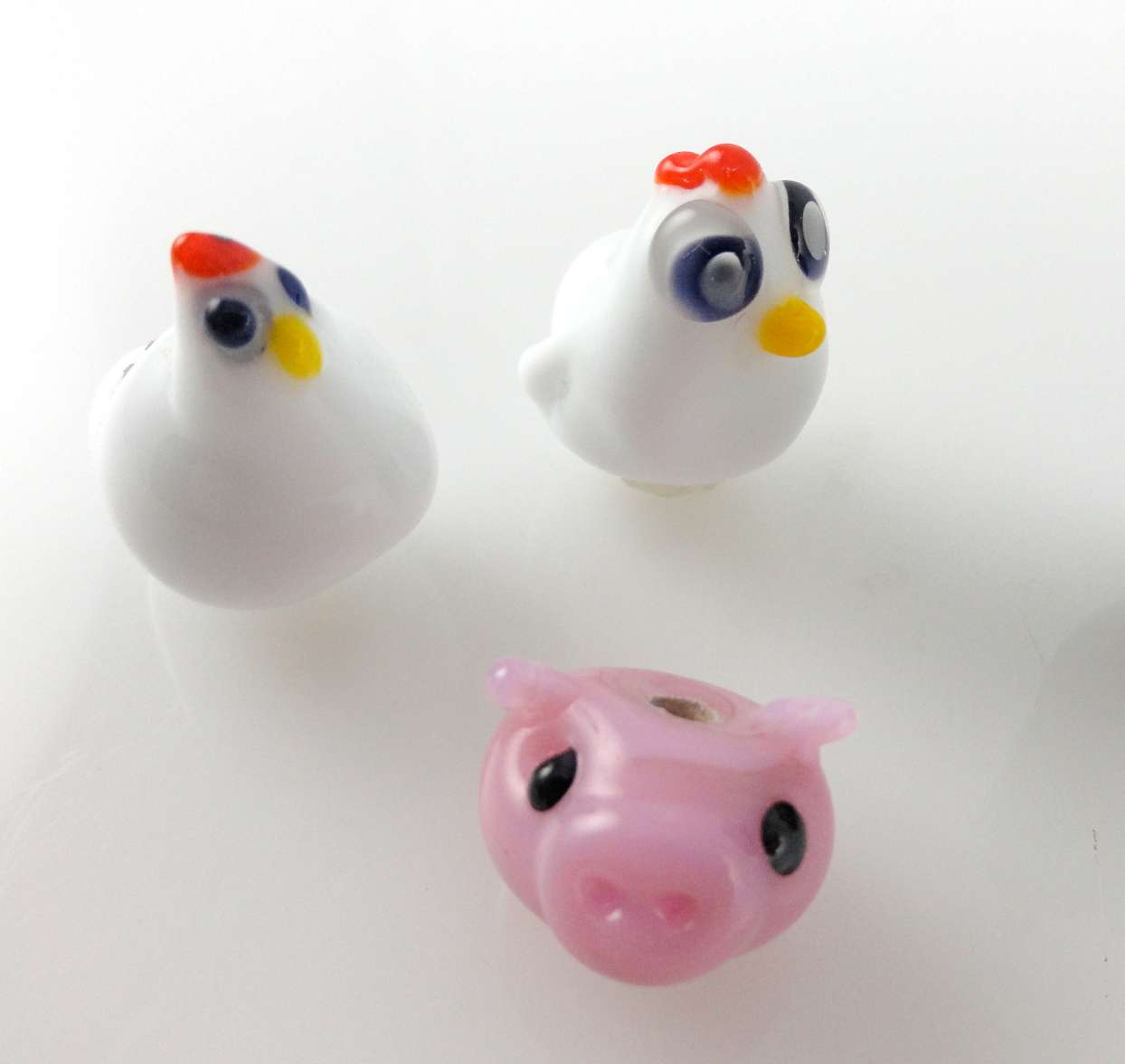 three sculptural glass beads. Two are shaped like chickens and one like the face of a pig